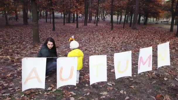 Mãe com crianças coletar folhas de outono no parque perto de uma guirlanda de folhas de papel branco com a inscrição Outono pintado com tintas coloridas . — Vídeo de Stock