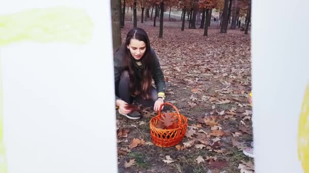 Мама с детьми собирает осенние листья в парке рядом с гирляндой из белых листьев бумаги с надписью Осень окрашена красочными красками . — стоковое видео