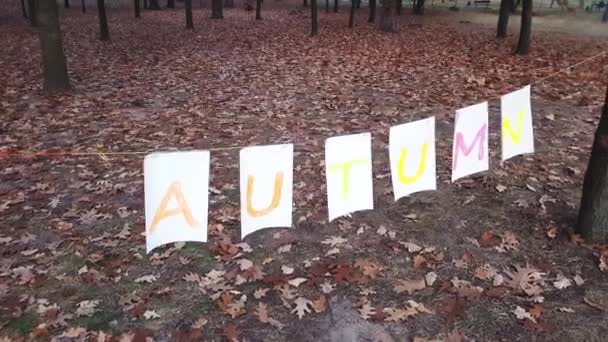 Mom with a small daughter hold a garland of white leaves of paper with the inscription Autumn in colorful autumn colors in the evening park. — Stockvideo