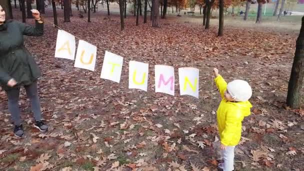 Küçük bir kızı olan anne, akşam parkında renkli sonbahar renkleriyle yazılmış beyaz yapraklı bir çelenk tutuyor.. — Stok video