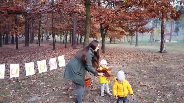 Η μαμά με τα παιδιά πετάνε φθινοπωρινά φύλλα στο πάρκο κοντά σε μια γιρλάντα από φύλλα λευκού χαρτιού με την επιγραφή Φθινόπωρο βαμμένα με πολύχρωμα χρώματα. — Αρχείο Βίντεο