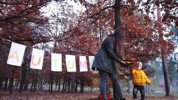 Mama z dziećmi podrzuca jesienne liście w parku przy girlandzie z białego papieru z napisem Jesień malowana kolorowymi farbami. — Wideo stockowe