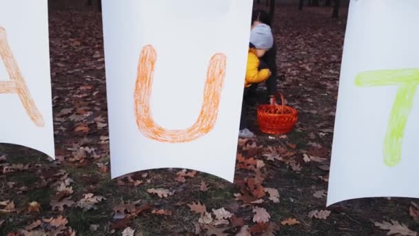 Μαμά με τα παιδιά συλλέγουν φύλλα του φθινοπώρου στο πάρκο κοντά σε μια γιρλάντα από φύλλα λευκού χαρτιού με την επιγραφή Φθινόπωρο βαμμένα με πολύχρωμα χρώματα. — Αρχείο Βίντεο