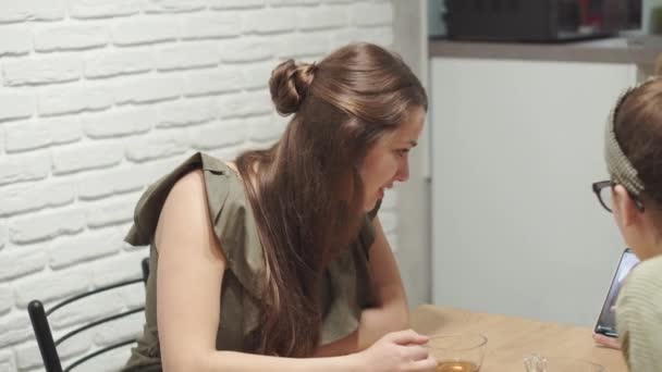 Cafés e restaurantes, homeliness, comunicação e conceito de tecnologia duas jovens irmãs conversando beber chá sentado em vestidos na mesa da cozinha e olhando para smartphone em casa à noite — Vídeo de Stock