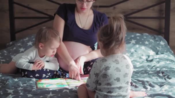 Terhesség, anyaság, család, gyermekkor, nevelés, képzés, elszigeteltség koncepciója - egy fiatal terhes anya kisgyermekekkel az ágyon könyvet olvas. Fiú és lány játszanak anyával otthon. — Stock videók