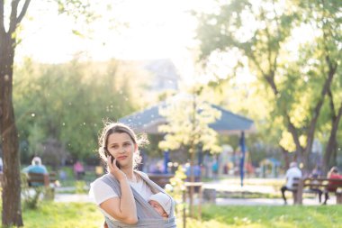 Annelik, bakım, bebekler, yaz, iletişim, ebeveynlik konsepti - Yeni doğmuş bebeğiyle parkta gün batımının arka ışığında yürüyen ve telefonla konuşan genç güzel anne.