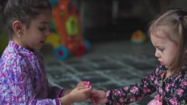 Çocukluk, oyunlar, mücevherler. Farklı milletlerden mor ve pembe elbiseli iki küçük kız yere oturmuş bilezik takıyor.