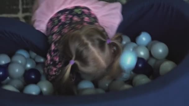 Childhood, games, toy, development, entertainment concept - Little three-year-old girl in pink dress play in dry bacein with multicolored ball lies down and howsle as if swimming in childrens room — Stock Video