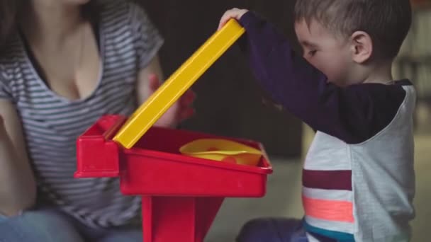 La maternidad, la infancia, la alimentación, la comida, la familia, el concepto de cuidado: la mamá de cabello oscuro de plan medio alimenta a su hijo de dos años de edad con apariencia de Oriente Medio desde el plato de cuchara en vaqueros rasgados sentados en el suelo — Vídeo de stock