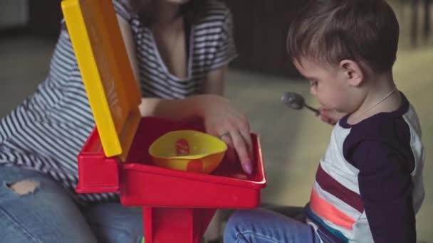 La maternidad, la infancia, la alimentación, la comida, la familia, el concepto de cuidado: la mamá de cabello oscuro de plan medio alimenta a su hijo de dos años de edad con apariencia de Oriente Medio desde el plato de cuchara en vaqueros rasgados sentados en el suelo — Vídeos de Stock