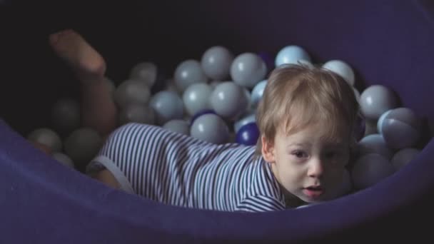 Childhood, games, toy, development, entertainment concept - little two-year-old boy in striped bodysuit play in dry bacein with multicolored ball lies down and howsle as if swimming in childrens room — Stock Video