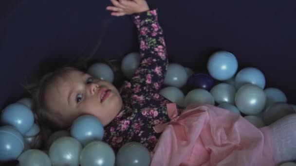 Детство, игры, игрушки, развитие, развлекательная концепция - Маленькая трехлетняя девочка в розовом платье играет в сухой бекон с разноцветным мячом лежит и воет, как будто плавает в детской комнате — стоковое видео