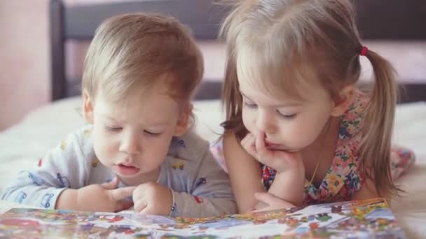 Niños encantadores - hermano y hermana, leyendo un libro, en la cama. Primer plano de los niños en la cama leyendo un libro. Un niño y una niña de blanco están jugando en la cama. Bebés de blanco en la cama. Lindos niños . — Vídeos de Stock