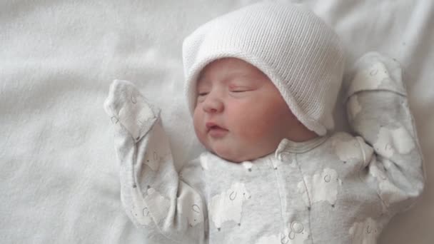 Niemowlę, dzieciństwo, rozwój, medycyna i koncepcja zdrowotna - widok z bliska z góry twarzy noworodka spokojne dziecko ubrane w body suit i kapelusz grimaces leżące na plecach na białym beżowym tle — Wideo stockowe