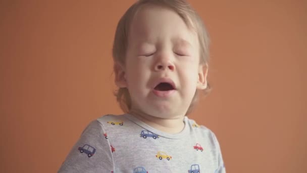 Děti, oslavy, narozeniny, herní koncept - zblízka malý blonďatý chlapec v šedém teplákovém obleku kýchne s úsměvem při pohledu do kamery a ukazuje různé emoce na oranžovém pevném pozadí. — Stock video