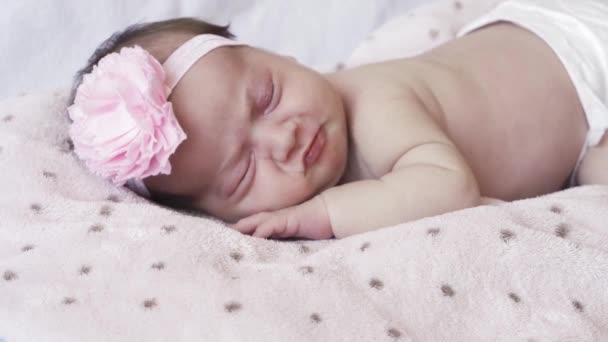 Βρεφική ηλικία, παιδική ηλικία, ανάπτυξη, ιατρική και υγειονομική έννοια - γκρο πλαν πρόσωπο ενός νεογέννητου γυμνού κοριτσιού που κοιμάται ξαπλωμένη στο στομάχι της με έναν επίδεσμο και ένα λουλούδι στο κεφάλι της σε ροζ φόντο. — Αρχείο Βίντεο