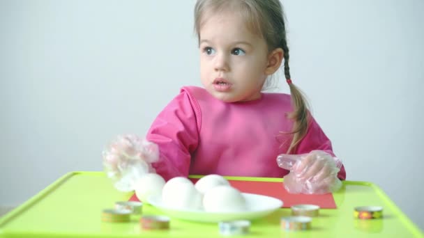 Paskalya, Hamursuz Bayramı, çocukluk, yaratıcılık, oyun konsepti - Sloven beyaz önlüklü üç yaşındaki tatlı sarışın kızın yakın çekimi plastik eldivenle Paskalya yumurtalarını boyuyor. — Stok video