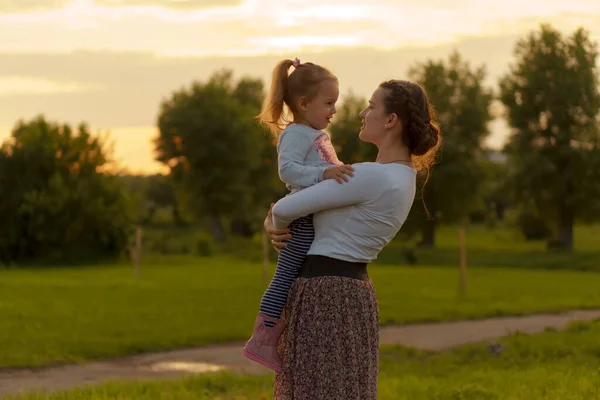 Anyaság, gondozás, csecsemők, nyár, szülői koncepció - Fiatal anya három éves kislány lánya a karjában sétál, és néz a gyermek hátterében naplemente a parkban közel virágzó bokor. — Stock Fotó