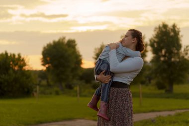 Annelik, bakım, bebekler, yaz, ebeveynlik konsepti - 3 yaşında küçük kızı olan genç bir anne kollarında yürüyor ve gün batımında çiçek açan çalıların yanındaki parkta çocuğa bakıyor.