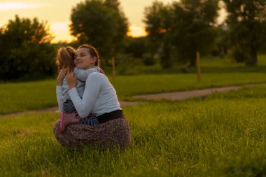 Annelik, bakım, bebekler, yaz, ebeveynlik konsepti - 3 yaşında küçük kızı olan genç bir anne kollarında yürüyor ve gün batımında çiçek açan çalıların yanındaki parkta çocuğa bakıyor.