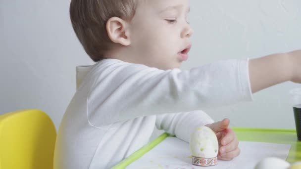 Egy kis szőke fiú tojást fest a kajafestéssel. Húsvéti előkészületek. Kéz a kézben. Kreatív fejlesztés 3 év alatti gyerekeknek. Oktatás, rajzolás otthon a karantén alatt — Stock videók
