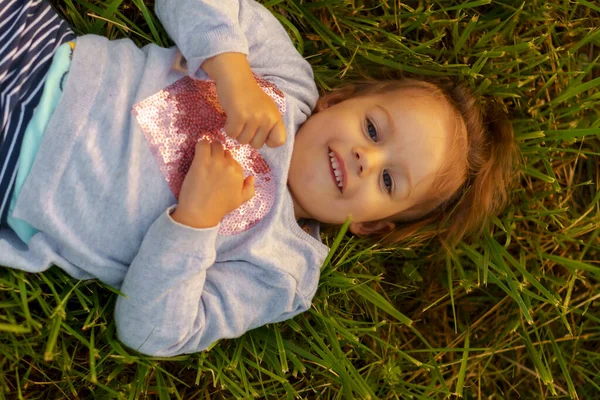 Infância, verão, lazer e entretenimento conceito close-up vista superior de um pouco bonito menina caucasiana de três anos de idade loira deitado na grama e sorrindo nos raios de pôr do sol — Fotografia de Stock