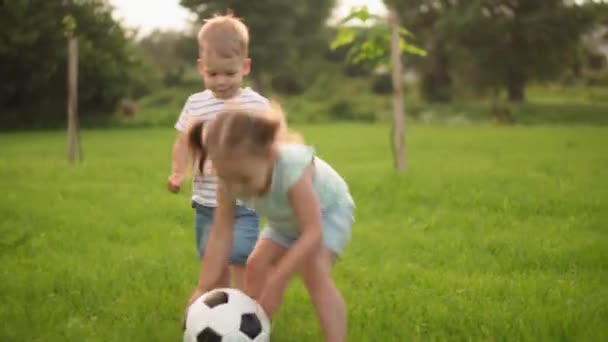 Infância, jogos e entretenimento, esportes, cultura física, parques e conceito ao ar livre - meninos e meninas crianças jogam futebol com bola preta e branca no gramado verde brilhante no parque ao pôr do sol. — Vídeo de Stock