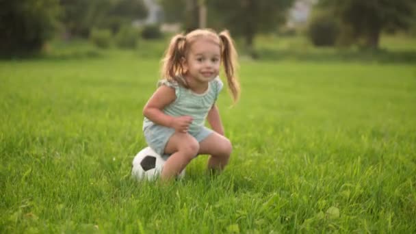 Dzieciństwo, gry i rozrywka, sport, koncepcja kultury fizycznej - małe dziewczynki w spodenkach i dwa kucyki grać siedzi na piłce nożnej czarno-białe piłkę na jasnozielonym trawniku w parku o zachodzie słońca — Wideo stockowe