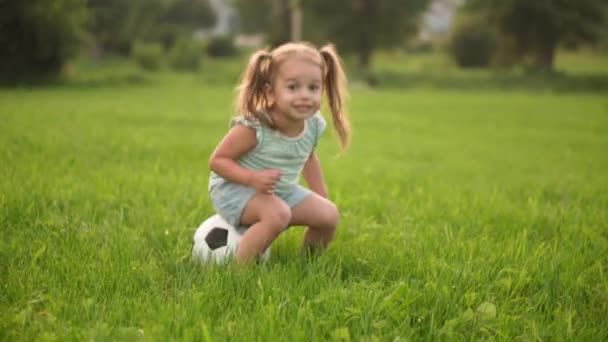 Infância, jogos e entretenimento, esportes, conceito de cultura física meninas em shorts e dois rabo de cavalo jogar sentado no futebol bola preta e branca no gramado verde brilhante no parque ao pôr do sol — Vídeo de Stock