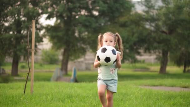 Infancia, juegos y entretenimiento, deportes, cultura física, concepto al aire libre - niñas en pantalones cortos y dos colas de caballo juegan al fútbol con bola blanca y negra en el césped verde brillante en el parque al atardecer — Vídeos de Stock