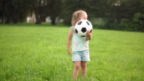 Dzieciństwo, gry i rozrywka, sport, kultura fizyczna, koncepcja plenerowa - małe dziewczynki w spodenkach i dwa kucyki grają w piłkę nożną czarno-białą na jasnozielonym trawniku w parku o zachodzie słońca — Wideo stockowe