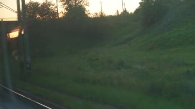 Ulaşım, seyahat, yol, demiryolu, manzara, çiftleşme konsepti - yaz aylarında günbatımından önce çayırların ve ormanların manzarasında cam ve paralel raylar üzerinde parlayan hız treninin penceresinden görünüm