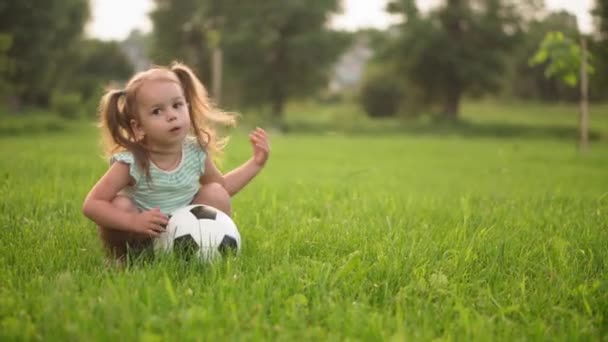Kindheit, Spiel und Unterhaltung, Sport, Körperkultur, Open-Air-Konzept - kleine Mädchen in kurzen Hosen und zwei Pferdeschwänzen spielen Fußball mit schwarz-weißem Ball auf hellgrünem Rasen im Park bei Sonnenuntergang — Stockvideo