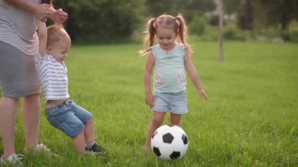 Dzieciństwo, gry i rozrywka, sport, kultura fizyczna, parki i koncepcja pleneru - mali chłopcy i dziewczęta z mamą grają w piłkę nożną czarno-białą na zielonym trawniku w parku o zachodzie słońca. — Wideo stockowe