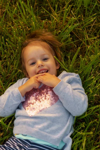 Kindheit, Sommer, Freizeit- und Unterhaltungskonzept - Nahaufnahme eines kleinen süßen dreijährigen blondhaarigen kaukasischen Mädchens, das im Gras liegt und in den Sonnenstrahlen lächelt — Stockfoto