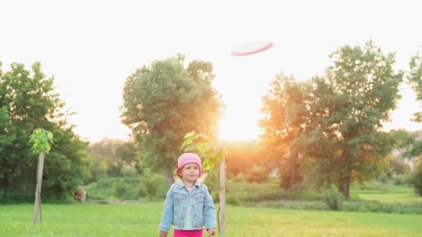Barndom, spel och underhållning, sport, fysisk kultur, parker och utomhus koncept - små barn flickor i crimson byxor och panama leka med flygande skiva på ljus grön gräsmatta i parken vid solnedgången. — Stockvideo