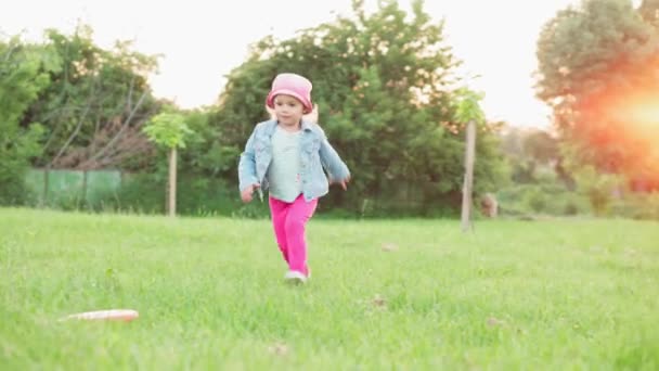 Infancia, juegos y entretenimiento, deportes, cultura física, parques y concepto al aire libre - niñas niños pequeños en pantalones carmesí y panama juegan con disco volador en el césped verde brillante en el parque al atardecer. — Vídeo de stock