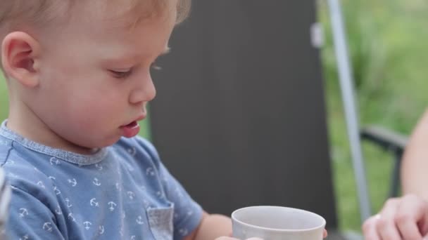 大きな家族、子供時代、食べ物、夏の休暇のコンセプト:青いTシャツの小さなブロンドの男の子のクローズアップ日陰で晴れた暑い日に屋外の庭のカントリーハウスのコテージでのピクニックのカップからの飲み物. — ストック動画