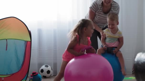 Barndom, Moderskap, spel, uppfostran och utveckling koncept - allmän plan för små barn pojke och flicka i samma ålder med ung mamma är glatt hoppa på rosa fitball och le i lekrum — Stockvideo