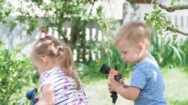 Colheita de verão, infância, conceito de alimentação saudável - plano geral de crianças pequenas, menino e menina, gêmeos irlandeses de 2 e 3 anos, estão derramando água de mangueiras em plantas no jardim da casa de campo. — Vídeo de Stock