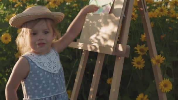 Dzieciństwo, koncepcja stylu prowansalskiego - mała 3-letnia blondynka w niebieskiej sukience i słomkowym kapeluszu rysuje się na sztaludze w pobliżu krzaków żółtych chryzantemy lawendowe pole w letni wieczór przed zachodem słońca — Wideo stockowe