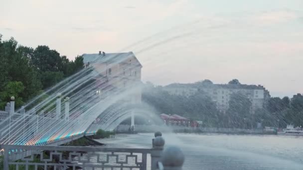 Landschaft, Teiche, Parks, Sommererholungskonzept - bunte Springbrunnen mit Beleuchtung in Form eines Bogens, der am Abend vor Sonnenuntergang vom Ufer in den großen See fällt im Parkbereich der Stadt in der Nähe des Hafens — Stockvideo