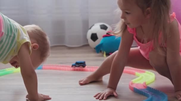 Jeugd, spelletjes, opvoeding en ontwikkeling concept - algemeen plan van kleine kinderen jongen en meisje van dezelfde leeftijd spelen met de constructeur van racebaan met speelgoed gemotoriseerde auto in de speelkamer van kinderen. — Stockvideo