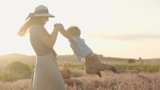 Große Familie, Vaterschaft, Kindheit, Mutterschaft, Provence-Stil Konzept - Zeitlupe junge große Mutter im Kleid schwingt kleinen Jungen an den Armen auf Lavendelfeld Blume am Sommerabend vor Sonnenuntergang — Stockvideo