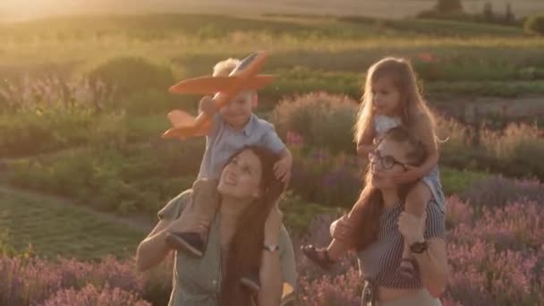 Grande família, infância, maternidade, conceito de estilo provence - Duas jovens mães felizes com crianças nos ombros menino e menina segurando e movendo avião no campo de lavanda na noite de verão antes do pôr do sol — Vídeo de Stock