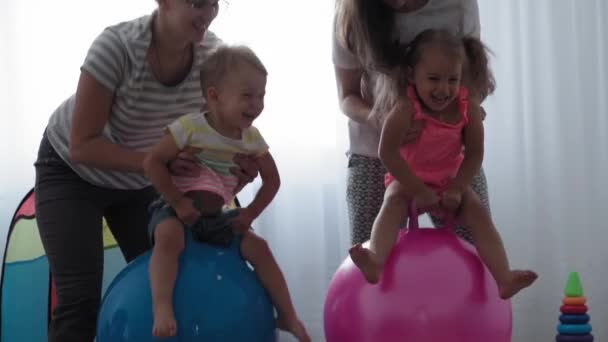 Dzieciństwo, macierzyństwo, gry, wychowanie i rozwój koncepcja - ogólny plan małych dzieci chłopiec i dziewczyna w tym samym wieku z młodą mamą są radośnie skaczą na różowym fitball i uśmiech w pokoju zabaw dla dzieci — Wideo stockowe