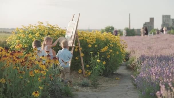 Gyermekkor, provence stílus koncepció - kis 3 gyerek fiú és szőke kislány kék ruhában és szalma kalap felhívja festőállvány közelében bokor sárga krizantém levendula mező nyáron este naplemente előtt — Stock videók