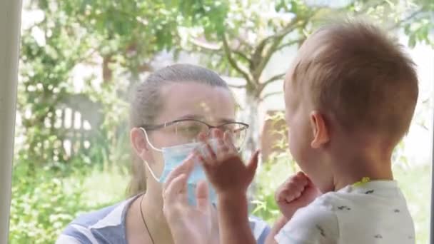 La mamma con un bambino piccolo con una maschera medica guarda fuori dalla finestra durante la quarantena a causa del coronavirus, covid-19 minaccia di virus. Auto isolamento. Primo piano. — Video Stock