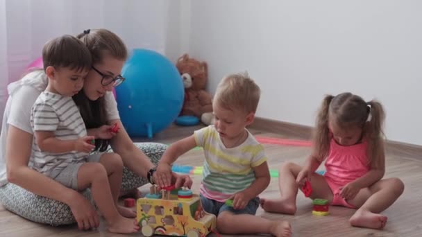 Infancia, juegos, crianza y concepto de desarrollo - plan general de los niños pequeños niño y niña de la misma edad con mamá están jugando con los niños constructor de coches de madera y rompecabezas en la sala de juegos para niños — Vídeo de stock