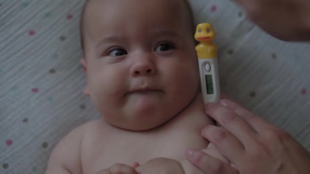 Säuglinge, Kindheit, Krankheit, Medizin und Gesundheit, Covid-19, Pandemie - Nahaufnahme weiblicher Hände misst Temperatur elektronisches Thermometer zu neugeborenen nackten Baby lächelt schief und weint auf dem Rücken liegend — Stockvideo